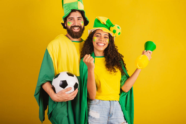 couple de supporters brésiliens de football, vêtus aux couleurs de la nation, femme noire, homme caucasien. utilisant le drapeau et les accessoires - Photo, image
