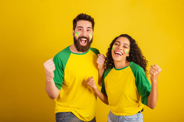 couple of brazil soccer supporters, dressed in the colors of the nation, black woman, caucasian man. Twisting and vibrating. - Fotografie, Obrázek