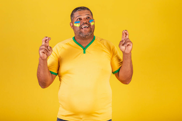 Brazilian black man, soccer fan from Brazil. Fingers crossed, wishing, sign of luck, cheering. - Фото, изображение