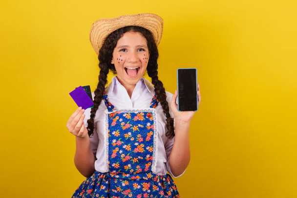 Girl wearing typical clothes for Festa Junina. holding cellphone and bank cards, apps, mobile shopping. For the Arraia party - Foto, Imagem