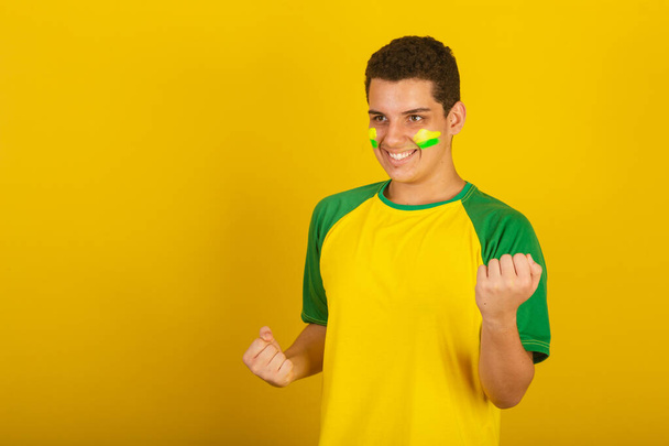 Fiatal brazil férfi focirajongó. zöld, sárga, csavaró és vibráló ruhába öltözve. - Fotó, kép