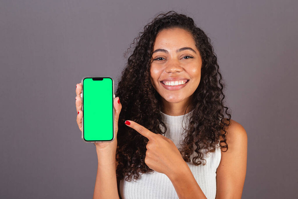 Young Brazilian black woman holding cellphone and showing screen, advertising photo. - Photo, image