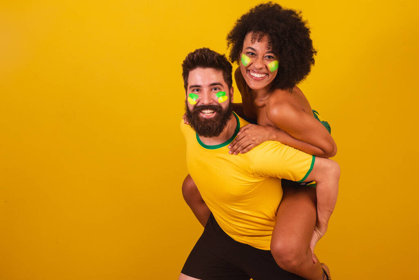 couple of brazilian soccer fans, dressed in the colors of brazil, black woman, caucasian man. joking, behind the back, in love - Foto, Imagen