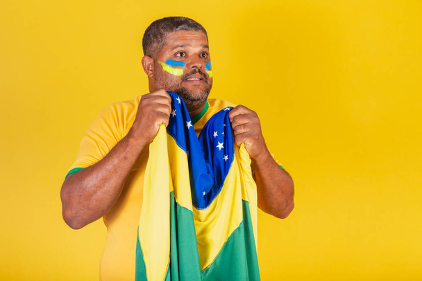 Brasilianischer Schwarzer, Fußballfan aus Brasilien. Besorgt, nervös, die brasilianische Fahne schwenkend. - Foto, Bild