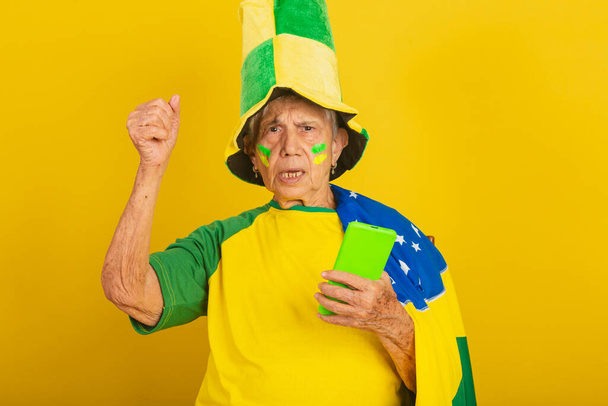 Elderly woman, soccer fan from Brazil, cheering with the flag. holding cellphone, smartphone - Valokuva, kuva
