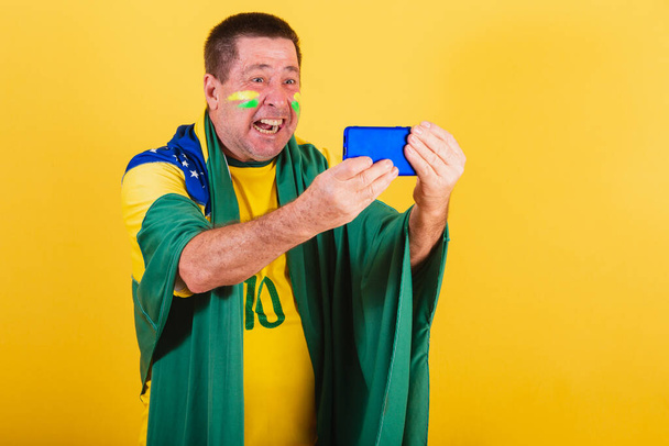 hombre adulto, aficionado al fútbol brasileño, utilizando la bandera, teléfono inteligente, viendo el juego por teléfono celular. en línea, streaming - Foto, imagen