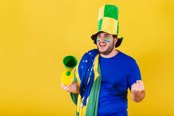 Brazylijski mężczyzna kaukaski, fan piłki nożnej z Brazylii, w kapeluszu i przy użyciu klaksonu, świętując zwycięstwo swojej drużyny.Cheering w mistrzostwach. - Zdjęcie, obraz