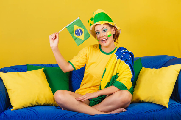 Caucasian woman, redhead, brazil soccer fan, brazilian, on couch holding brazil flag - Foto, immagini