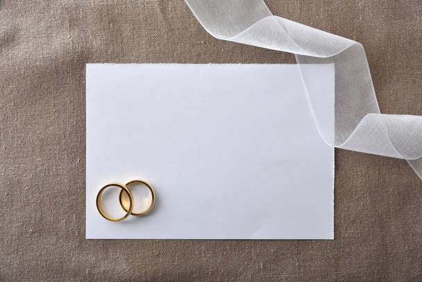 Hochzeitsankündigung Hintergrund mit goldenen Ringen im Papierausschnitt auf braunem Tuch und Detail der weißen Schleife. Ansicht von oben. - Foto, Bild