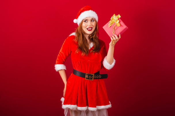 hermosa brasileña pelirroja, vestida con ropa de Navidad, Santa Claus. Dudas sobre lo que hay en este regalo rojo. - Foto, imagen
