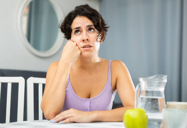 Dissatisfied young woman sitting at the table in the room of her apartment - Photo, image