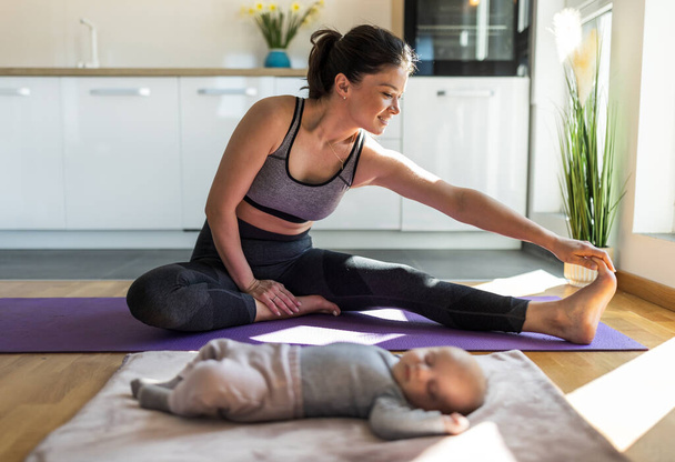 Jonge mooie moeder oefenen op yoga mat thuis in de voorkant van keukenkasten terwijl pasgeboren baby slapen naast haar. Gezonde geestelijke en lichamelijke conditie concept na de bevalling - Foto, afbeelding