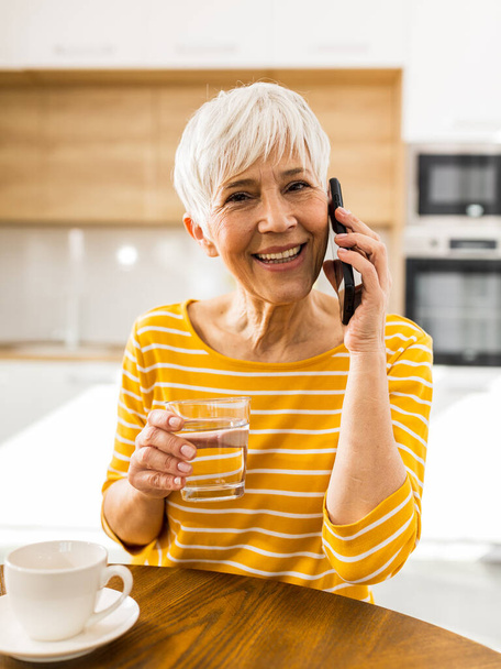 Melko kypsä nainen puhuu matkapuhelimella ja pitää lasin vettä ruokapöydässä kotona - Valokuva, kuva