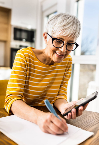 Улыбающаяся взрослая женщина, рассчитывающая счета за домашнее хозяйство на мобильный телефон и письменные расходы и сбережения за обеденным столом дома - Фото, изображение