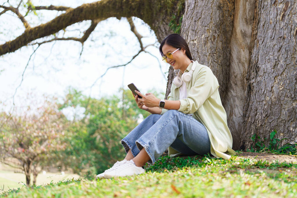 Donne sedute sull'erba per navigare sui social media sullo smartphone mentre relax sotto l'albero nel parco. - Foto, immagini