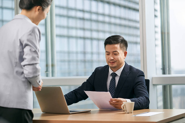 азиатский бизнес-менеджер рассматривает бумажный документ отчет, представленный ассистенткой в современном офисе - Фото, изображение