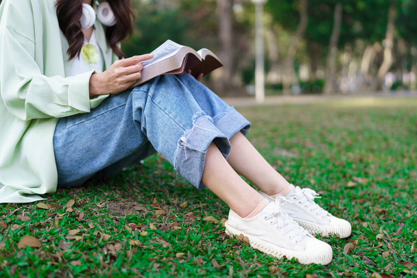 Mulheres em fone de ouvido em torno do pescoço e sentado na grama para ler um livro depois de andar de bicicleta no parque. - Foto, Imagem