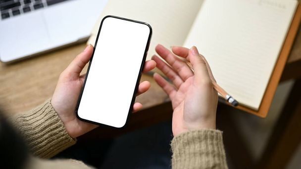Close-up shot of a woman in cozy sweater holding her smartphone, scrolling on her phone, or searching informations on the internet at her desk. phone white screen mockup - Zdjęcie, obraz