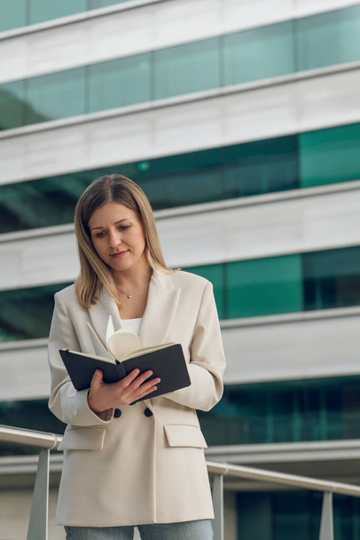 Confident young female entrepreneur in elegant outfit standing near modern office building while reading diary in city during work - Zdjęcie, obraz