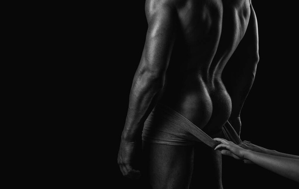 Female hand undresses pants. Bodybuilder and strip theme. Bare torso. Strong backside. Young mans naked ass and back on black isolated background. Metrosexual concept - Фото, зображення
