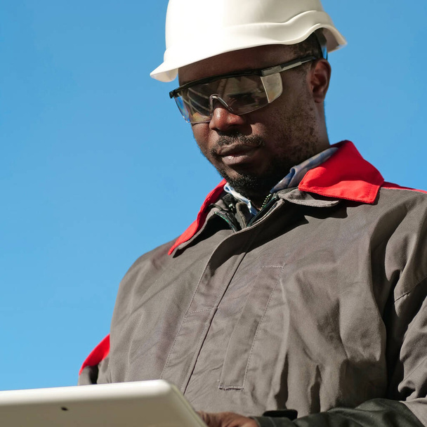 Afrikkalainen amerikkalainen työntekijä kypärä ja suojaava suojalasit tabletti tietokone seisoo rakennustyömaalla - Valokuva, kuva