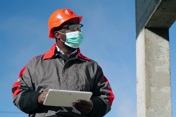 Αφροαμερικανός εργάτης με ιατρική προστατευτική μάσκα στέκεται στο εργοτάξιο με tablet υπολογιστή - Φωτογραφία, εικόνα
