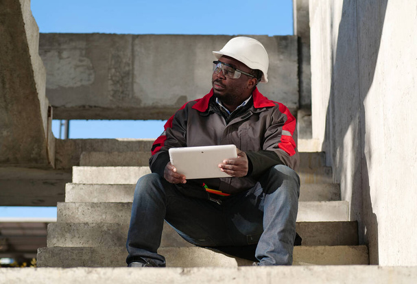 Trabajador afroamericano cansado con los ojos cerrados en blanco casco se sienta en el sitio de construcción y sostiene en las manos tableta PC - Foto, imagen