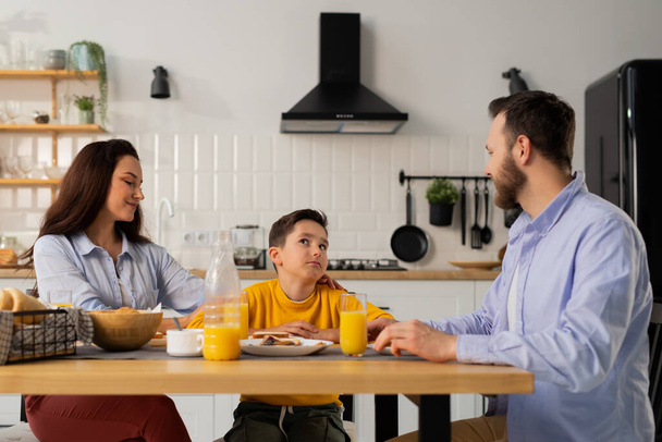 Ένα αγοράκι τρώει πρωινό με τους γονείς του στο τραπέζι της κουζίνας. Ένα αγόρι με κίτρινο μπουφάν επικοινωνεί με τους γονείς του κατά τη διάρκεια του πρωινού. Υψηλής ποιότητας φωτογραφία - Φωτογραφία, εικόνα