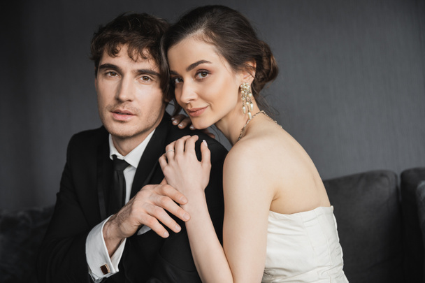 Porträt einer charmanten und jungen brünetten Braut in luxuriösen Ohrringen mit Perlen und weißem Hochzeitskleid, die ihren Bräutigam im schwarzen Anzug im Hotelzimmer umarmt  - Foto, Bild