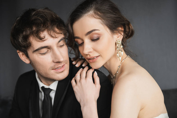 portrét krásné mladé nevěsty v luxusních náušnicích s perlami a náhrdelníkem sedí se zavřenýma očima a objímají šťastného ženicha v černém obleku v hotelovém pokoji - Fotografie, Obrázek