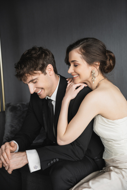 portrét půvabné mladé nevěsty v bílých svatebních šatech a pohledného ženicha v černém obleku, usmívající se společně v hotelovém pokoji, šťastné novomanžele - Fotografie, Obrázek