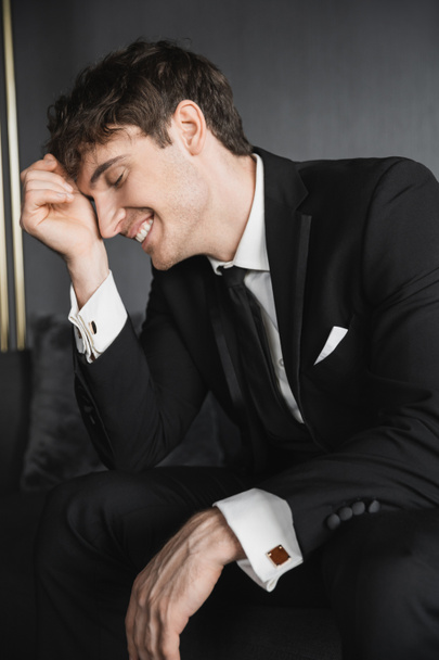 portrait of joyful and young groom in black suit with white shirt and tie touching face with hand while smiling and sitting on comfortable couch in modern hotel room on wedding day - Photo, Image