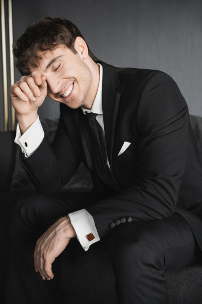 portrait of positive young groom in black suit with white shirt and tie touching face with hand while smiling and sitting on comfortable dark grey couch in modern hotel room on wedding day - Photo, Image