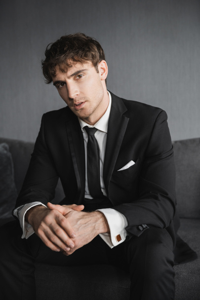 portrait of good looking young groom in black suit with white shirt and tie sitting on comfortable dark grey couch and looking at camera in modern hotel room on wedding day - Photo, Image