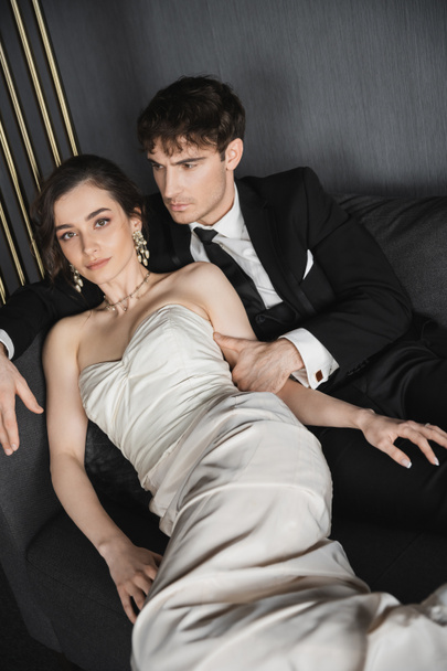 charming young bride in luxurious earrings with pearls and white wedding dress leaning on groom in black suit with tie while sitting together on dark grey couch in hotel room - Foto, Bild