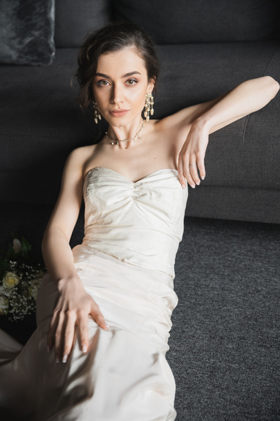 okouzlující mladá nevěsta s brunetkou vlasy sedí v elegantních a bílých svatebních šatech, luxusní šperky, náušnice a náhrdelník při pohledu do kamery v blízkosti svatební kytice v hotelovém pokoji  - Fotografie, Obrázek