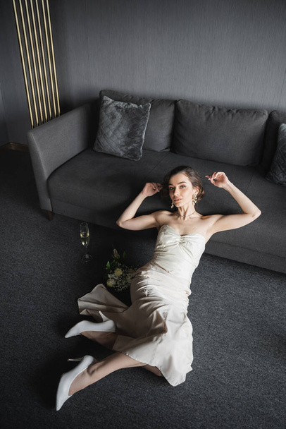 vysoký úhel pohledu nevěsty s brunetkou vlasy sedí v bílých svatebních šatech, vysoké podpatky a luxusní šperky vedle sklenice šampaňského a kytice na podlaze v blízkosti pohovky v hotelovém pokoji - Fotografie, Obrázek