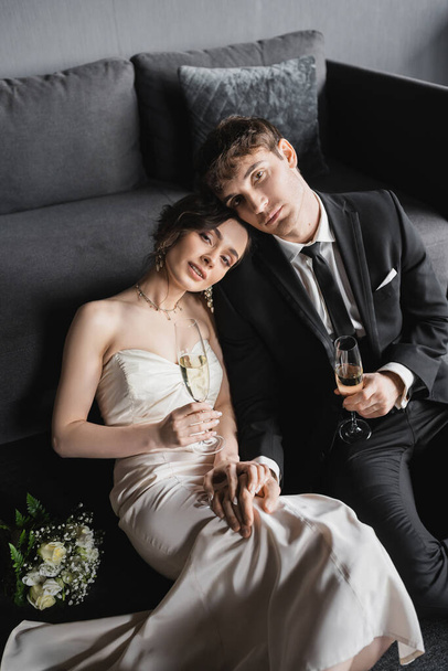 happy newlyweds, bride in white wedding dress and groom in black suit holding glasses of champagne while looking at camera after wedding ceremony and sitting near bridal bouquet in hotel room   - Fotografie, Obrázek