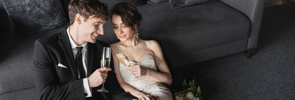 šťastná nevěsta v bílých svatebních šatech a ženich v černém obleku cinkající sklenice šampaňského při oslavě jejich manželství v blízkosti svatební kytice po svatbě v hotelovém pokoji, banner  - Fotografie, Obrázek