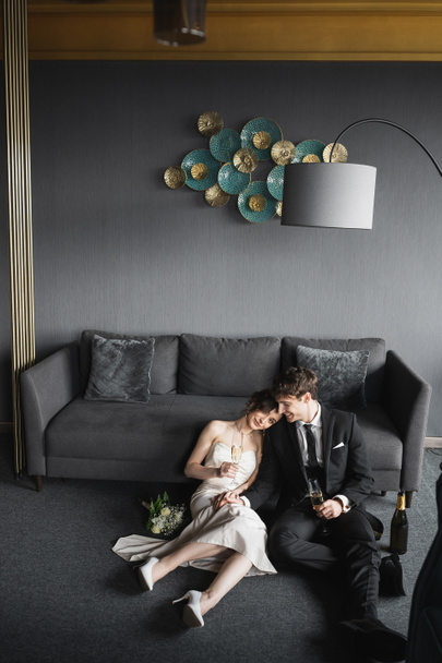 veselá nevěsta ve svatebních šatech opírající se o rameno ženicha v černém obleku a držící sklenice šampaňského v blízkosti svatební kytice, gauče a podlahové lampy v hotelovém pokoji  - Fotografie, Obrázek