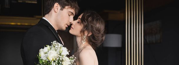 вид сбоку на красивого жениха в черной формальной одежде целоваться брюнетка невеста с роскошными украшениями держа свадебный букет цветов, стоя в вестибюле отеля, баннер  - Фото, изображение