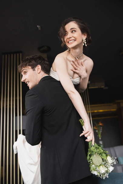 good looking groom in black formal wear lifting cheerful bride with makeup in white wedding dress holding bridal bouquet of flowers while standing in hotel lobby  - Foto, afbeelding