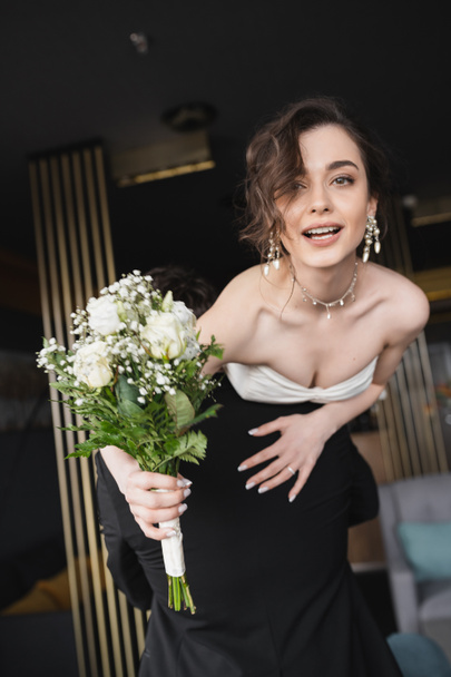 Bräutigam in schwarzer Festkleidung hebt hübsche Braut in weißem Hochzeitskleid und luxuriösem Schmuck und hält Brautstrauß mit Blumen in der Hotellobby  - Foto, Bild