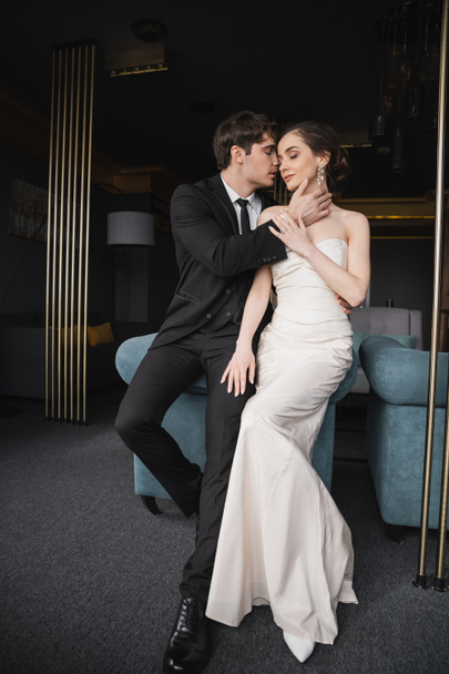tender groom in black suit with tie touching face of charming bride in white wedding dress and jewelry leaning on blue couch in modern hotel room - Photo, Image
