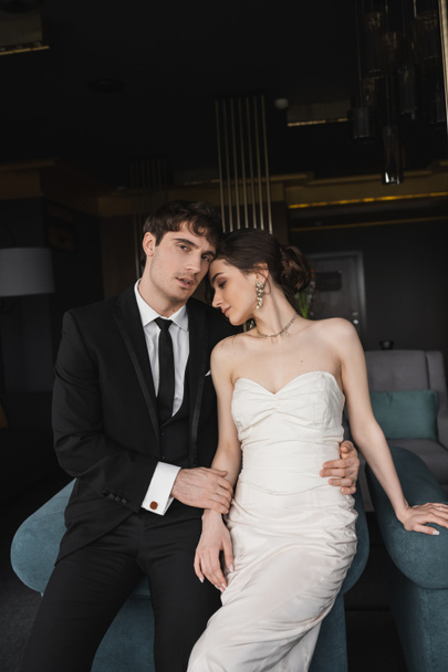 handsome groom in black suit looking at camera while hugging waist of charming bride in white dress and jewelry leaning on blue couch in modern hotel room  - Foto, afbeelding