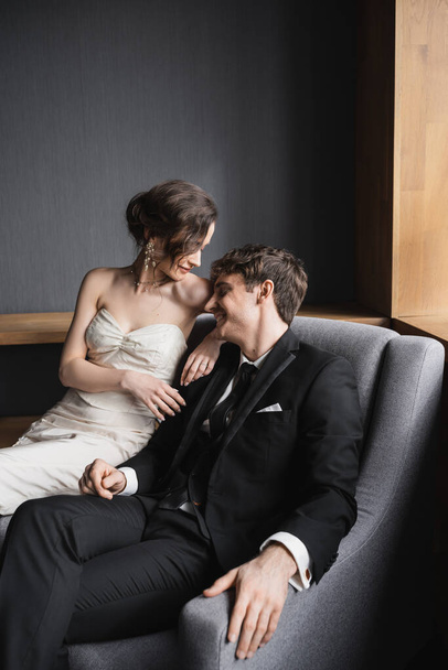 brunette bride in white wedding dress and luxurious jewelry sitting together with happy groom in black suit with tie on comfortable armchair in hotel room  - Foto, imagen
