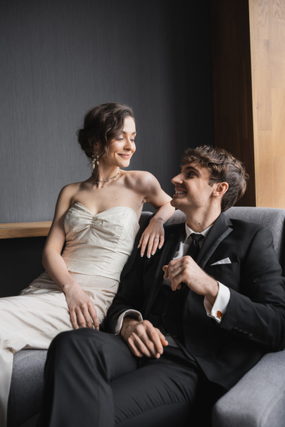 šťastná nevěsta v bílých svatebních šatech a luxusní náhrdelník s úsměvem při posezení spolu s pozitivním ženichem v černém obleku na pohodlném křesle v hotelovém pokoji  - Fotografie, Obrázek