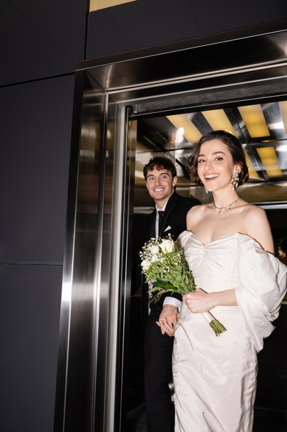 pretty bride in white dress holding bridal bouquet with flowers and hand of cheerful groom in suit walking out of elevator in hotel, happy newlyweds  - Photo, image