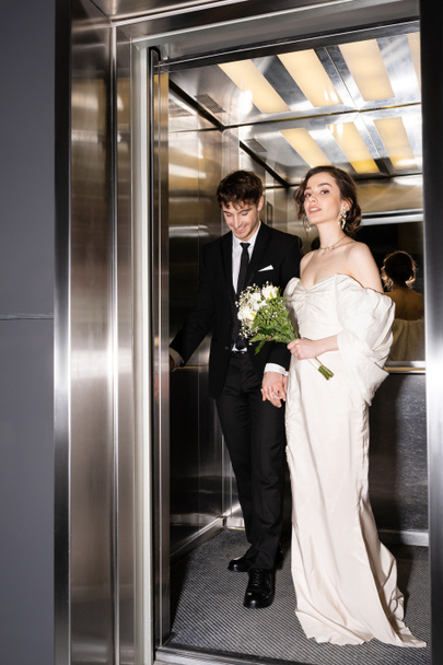 full length of pretty bride in white dress holding bridal bouquet with flowers and hand of cheerful groom in suit standing in elevator in hotel, happy newlyweds  - Fotó, kép