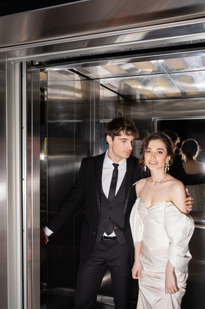 gut aussehender Bräutigam in formeller Kleidung, der Knopf drückt und fröhliche Braut im Brautkleid umarmt, während sie zusammen im Fahrstuhl des modernen Hotels steht  - Foto, Bild